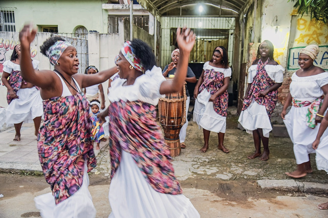 Imagem: Fotografia. No centro, duas mulheres negras estão dançando. Em volta delas, mais mulheres negras estão sorrindo e uma delas está com as mãos sobre um atabaque. Todas estão com faixa colorida na cabeça, vestido branco e tecido colorido em volta do corpo Fim da imagem.