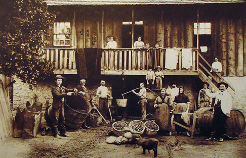 Imagem: Fotografia em sépia. No centro, homens com chapéu e ternos estão olhando para frente e entre eles há vários barris, cestos e duas mulheres com vestidos. Ao fundo há uma mulher e crianças na varanda de uma casa. Fim da imagem.