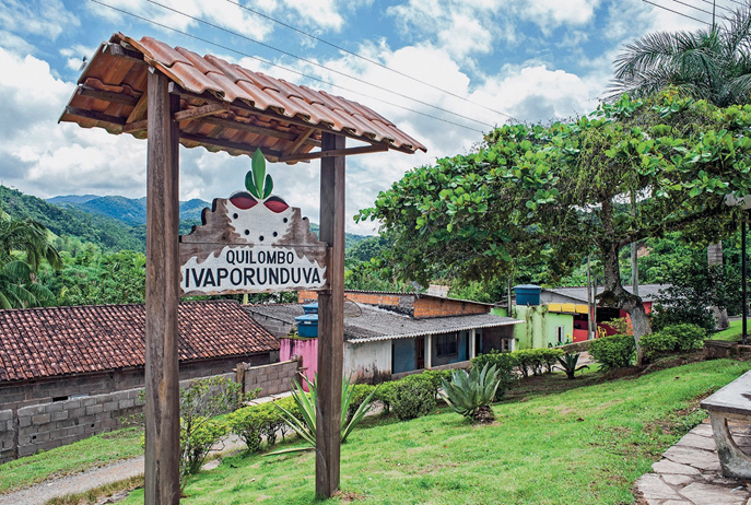 Imagem: Fotografia. No centro, placa com a informação: Quilombo Ivaporunduva. Ao fundo, casas, árvores e morros. Fim da imagem.