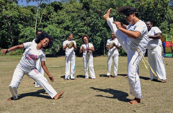 Imagem: Fotografia. Duas pessoas estão sorrindo e lutando capoeira. Atrás, cinco pessoas estão segurando instrumentos musicais. Todos estão com roupas brancas e descalços. Ao fundo, árvores. Fim da imagem.
