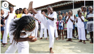 Imagem: Fotografia c. Duas crianças estão lutando capoeira. Em volta delas, várias crianças estão sorrindo e batendo palmas. Todas estão com roupas brancas.  Fim da imagem.