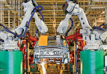 Imagem: Fotografia. Dois braços mecânicos ao lado de uma estrutura de carro. Ao fundo, máquinas. Fim da imagem.