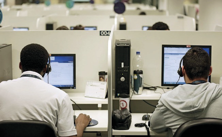 Imagem: Fotografia. Dois homens estão com fones de ouvido sobre as cabeças e sentados atrás de mesas com computadores em cima. Entre eles há uma divisória e ao fundo, mais pessoas sentadas atrás de computadores. Fim da imagem.