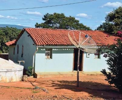 Imagem: Fotografia. Uma antena parabólica fincada no chão de terra. Atrás há uma casa e árvores. Fim da imagem.