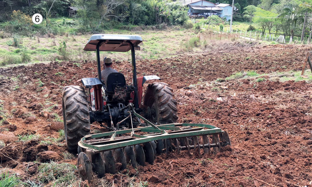 Imagem: Fotografia 6. Um homem está dirigindo um trator sobre um terreno plano com terra. Ao fundo, plantas e árvores.  Fim da imagem.