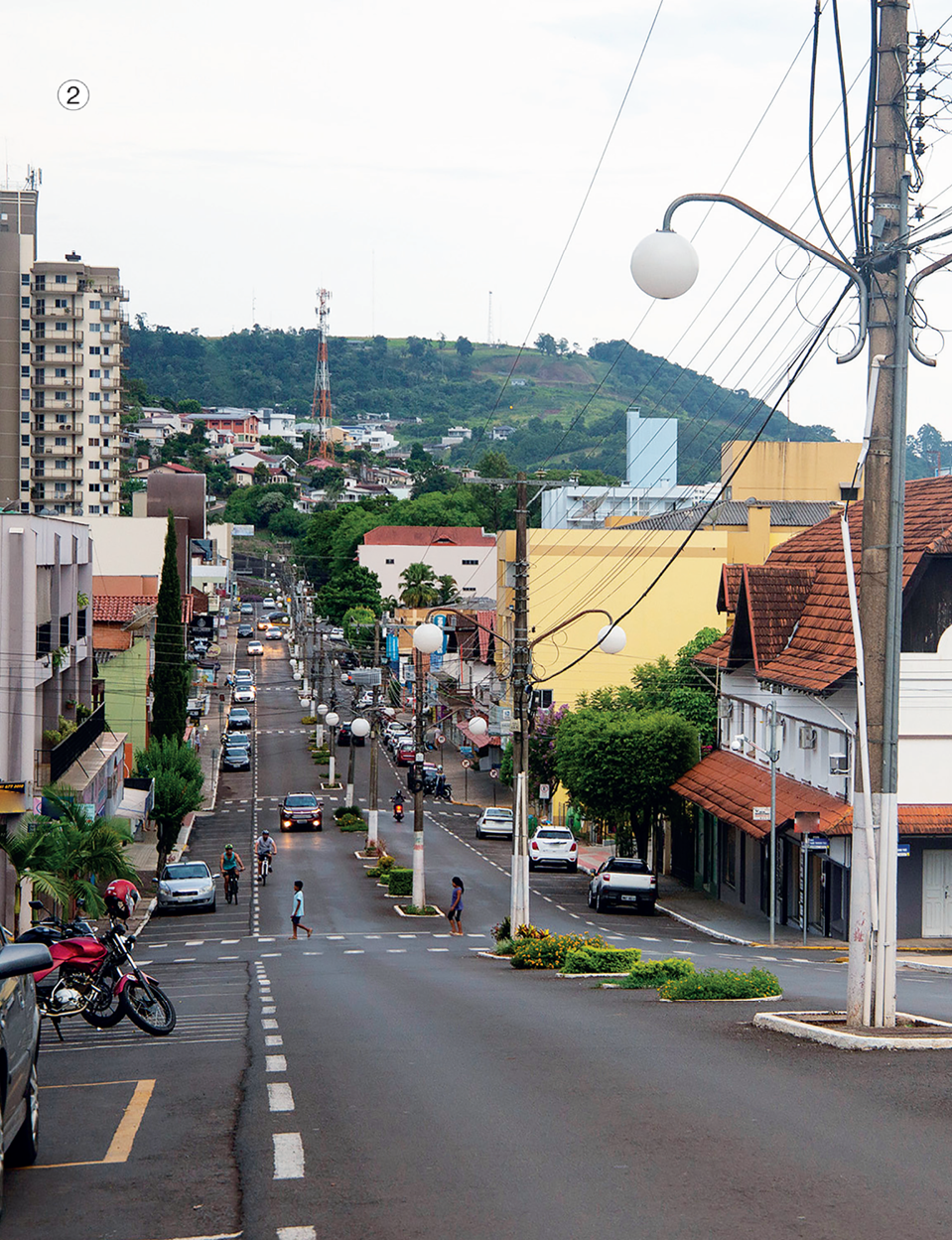 Imagem: Fotografia 2. No centro, pessoas estão atravessando uma faixa de pedestres. Atrás há carros sobre uma rua asfaltada. Nas laterais há postes, árvores e casas. Ao fundo, prédios, construções e um morro com árvores. Fim da imagem.