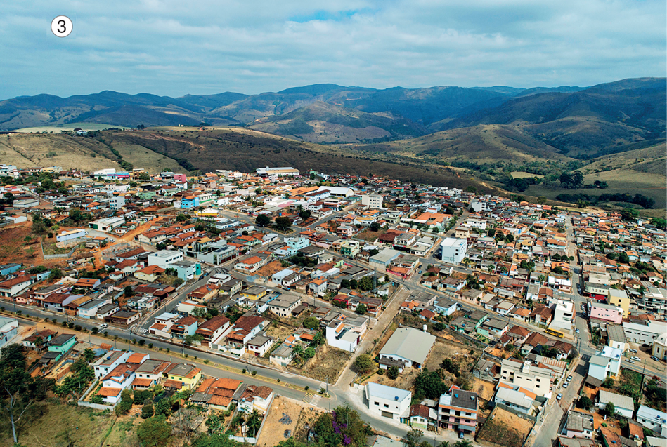 Imagem: Fotografia 3. Vista aérea de ruas, casas e árvores. Ao fundo, morros.  Fim da imagem.