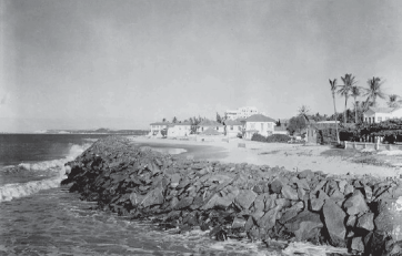 Imagem: Fotografia em preto e branco. À esquerda, o mar, à direita, um muro de pedras e ao fundo, areia, árvores e algumas casas.   Fim da imagem.