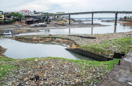 Imagem: Fotografia. Um rio com muito lixo e água escura. Ao fundo, uma ponte e construções.   Fim da imagem.