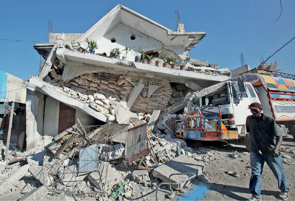 Imagem: Fotografia. Um homem está andando ao lado de uma casa, que está caída sobre destroços.  Fim da imagem.