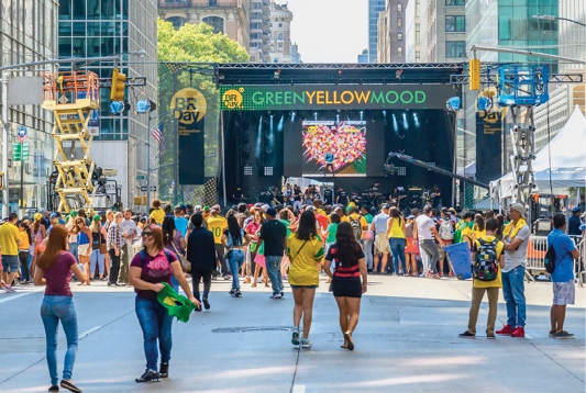 Imagem: Fotografia. Várias pessoas na frente de um palco. Acima há uma faixa com a informação: GREEN YELLOW MOOD. Ao fundo, prédios.  Fim da imagem.