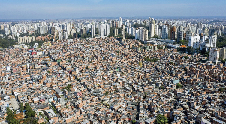 Imagem: Fotografia. Vista aérea de uma cidade com várias casas simples e árvores. Ao fundo, vários prédios e casas grandes. Fim da imagem.