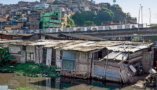 Imagem: Fotografia. Construções de madeira sobre água escura. Ao fundo, uma favela sobre um morro e árvores. Fim da imagem.