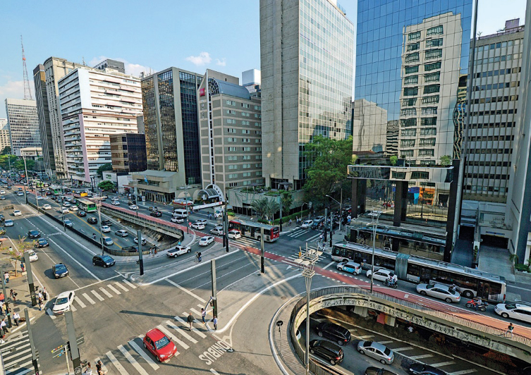 Imagem: Fotografia. Vista aérea de automóveis sobre ruas asfaltadas. Nas laterais há vários prédios e algumas árvores. Fim da imagem.