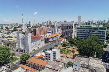 Imagem: Fotografia. Vista aérea de uma praça pequena com árvores. Em volta há prédios, construções, ruas e árvores.  Fim da imagem.