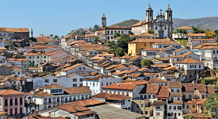 Imagem: Fotografia. Vista aérea de várias casas aglomeradas. Ao fundo, uma igreja sobre um morro.   Fim da imagem.