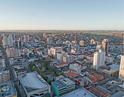 Imagem: Fotografia. Vista aérea de prédios, casas, ruas e árvores.   Fim da imagem.