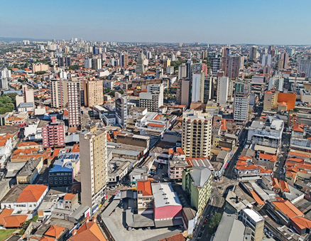 Imagem: Fotografia. Vista aérea de prédios, casas, ruas e árvores.   Fim da imagem.