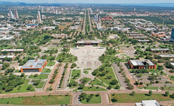 Imagem: Fotografia. Vista aérea de uma praça grande e extensa com várias árvores. Em volta há ruas, construções e casas. Ao fundo, alguns prédios.   Fim da imagem.