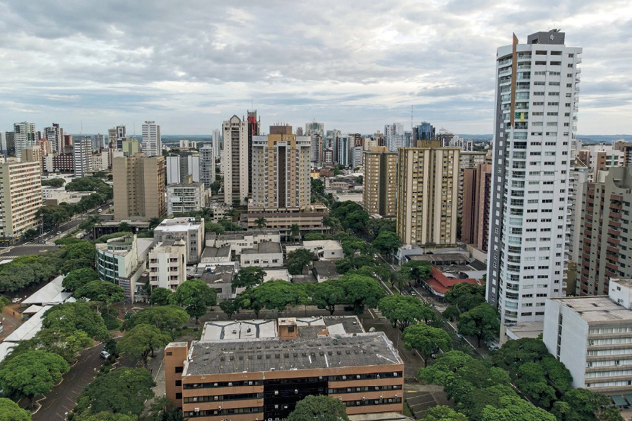 Imagem: Fotografia. Vista aérea de ruas com árvores em volta, prédios e casas.  Fim da imagem.