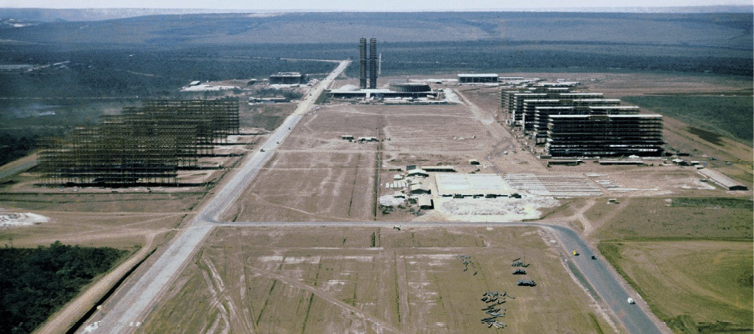 Imagem: Fotografia. Terreno plano e seco. No centro, construção de duas torres e nas laterais há duas ruas e construções de prédios. Ao fundo, morros.   Fim da imagem.