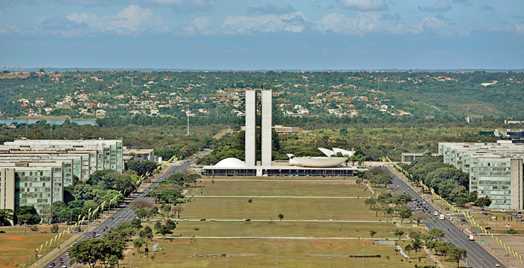 Imagem: Fotografia. Terreno verde com árvores. No centro há duas torres e dois semicírculos sobre construção extensa. Nas laterais há duas ruas asfaltadas e prédios. Ao fundo, casas e árvores sobre morros.  Fim da imagem.