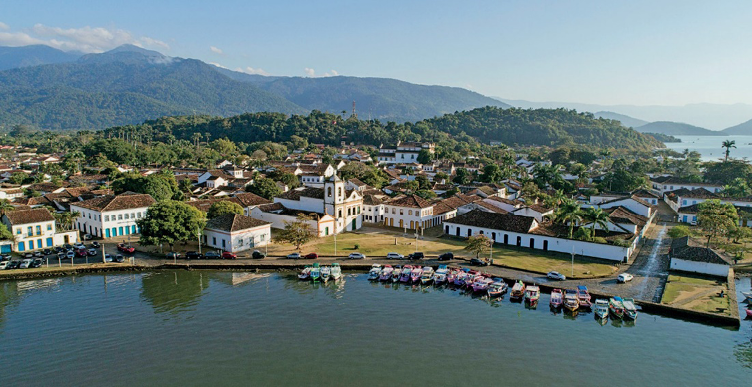 Imagem: Fotografia. Em primeiro plano há barcos atracados. Em segundo plano, casas e árvores. Ao fundo, morros e o mar.  Fim da imagem.