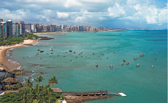 Imagem: Fotografia. Vista aérea de barcos no mar verde. À esquerda, praia, árvores e prédios.   Fim da imagem.