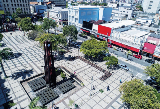 Imagem: Fotografia. Vista aérea de uma torre em uma praça com árvores em volta. Ao fundo, ruas e construções.  Fim da imagem.