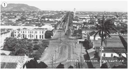 Imagem: Fotografia 1 em preto e branco. Vista aérea de uma rua de terra com postes enfileirados. Nas laterais há árvores e casas.   Fim da imagem.