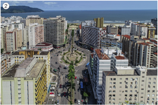 Imagem: Fotografia 2. Vista aérea de uma rotatória asfaltada. Em volta há vários prédios e ao fundo, o mar.  Fim da imagem.