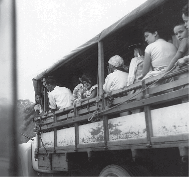 Imagem: Fotografia em preto e branco. Homens, mulheres e crianças sentadas na parte traseira de um caminhão.   Fim da imagem.