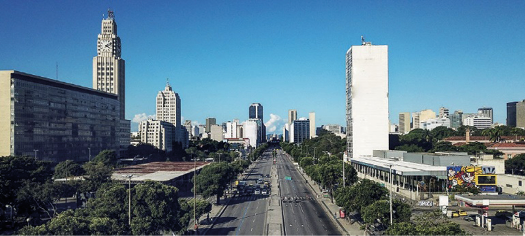 Imagem: Fotografia. No centro há duas avenidas asfaltadas. Nas laterais há árvores, prédios e construções. Fim da imagem.