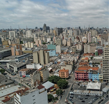 Imagem: Fotografia. Vista aérea de veículos nas ruas, prédios, casas e árvores.   Fim da imagem.