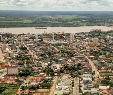 Imagem: Fotografia. Vista aérea de ruas, casas e árvores. Ao fundo, um lago grande e do outro lado, morros verdes.  Fim da imagem.