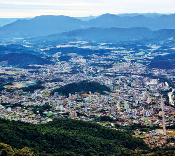 Imagem: Fotografia. Vista aérea de uma cidade ao lado de um morro verde. Ao fundo, mais morros.   Fim da imagem.