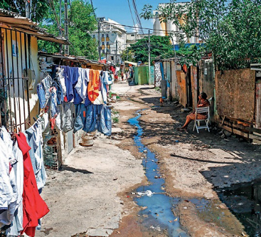 Imagem: Fotografia. No centro há uma poça d’água escura e extensa em uma rua. Nas laterais há casas improvisadas, árvores e roupas penduradas.   Fim da imagem.