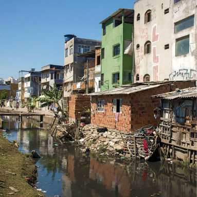 Imagem: Fotografia. No centro há um córrego com água escura. Ao lado há casas com três andares e árvores.  Fim da imagem.