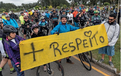 Imagem: Fotografia. Vários ciclistas com capacete de proteção estão segurando uma faixa com a informação: + RESPEITO. Ao fundo, árvores.  Fim da imagem.