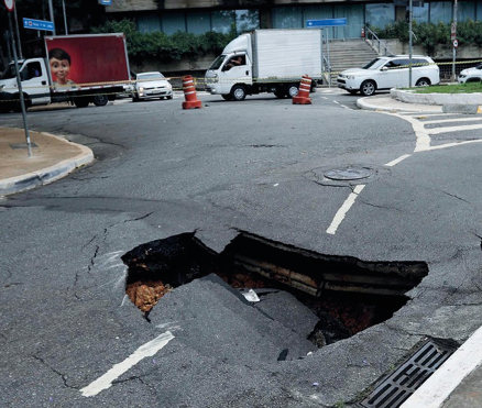 Imagem: Fotografia. Uma rua asfaltada com um buraco no meio. Ao fundo, cones de trânsito e carros enfileirados. Fim da imagem.