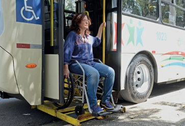 Imagem: Fotografia. Uma mulher em uma cadeira de rodas está descendo de um ônibus através de uma rampa na porta do veículo.   Fim da imagem.