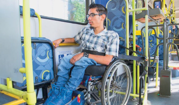 Imagem: Fotografia. Um jovem em uma cadeira de rodas está dentro de um ônibus. Atrás dele, a catraca.   Fim da imagem.