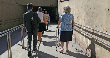 Imagem: Fotografia. Uma senhora está descendo uma rampa e se apoiando em um andador. Ao seu lado, pessoas estão descendo a rampa.  Fim da imagem.