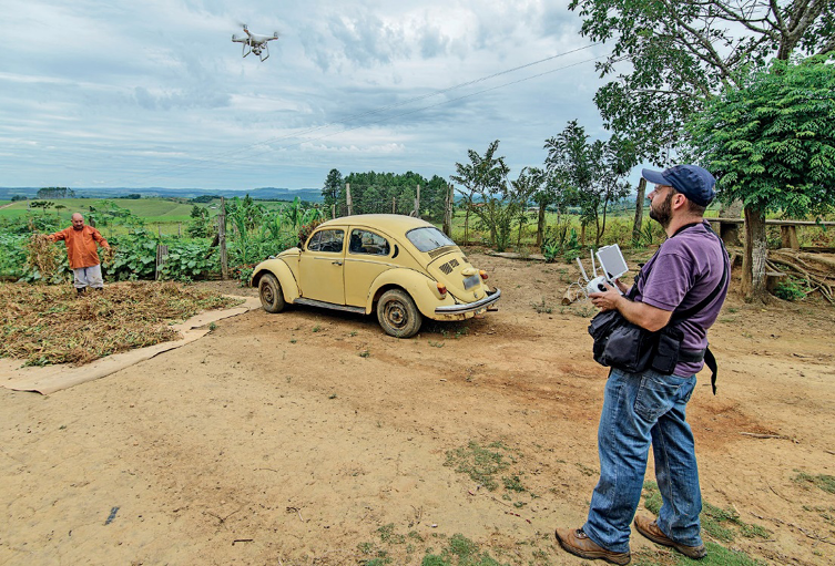 Imagem: Fotografia. Um homem está segurando um aparelho eletrônico e olhando para cima. Na frente dele há um drone voando sobre um fusca amarelo. Ao lado do carro há um homem sobre uma plantação. Ao fundo, árvores.  Fim da imagem.