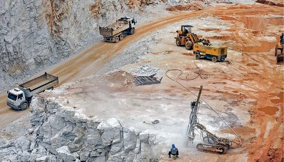 Imagem: Fotografia. Vista aérea de caminhões sobre terreno marrom e cinza. Ao lado há um morro cinza.  Fim da imagem.