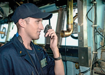 Imagem: Fotografia. Um homem com boné e uniforme azul-escuro está segurando um rádio na frente da boca. Ao fundo, janelas e cabos. Fim da imagem.
