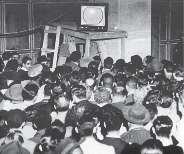 Imagem: Fotografia em preto e branco. Uma multidão na frente de uma televisão pequena, que está em cima de um suporte.  Fim da imagem.