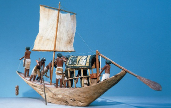 Imagem: Escultura de um barco com vela. Dentro há homens e tapeçarias. Ao lado há um remo grande pendurado.  Fim da imagem.
