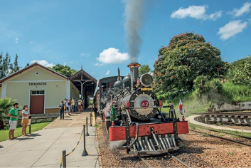 Imagem: Fotografia. Locomotiva expelindo fumaça sobre um trilho. Ao lado há pessoas e uma construção. Ao fundo, árvores.  Fim da imagem.