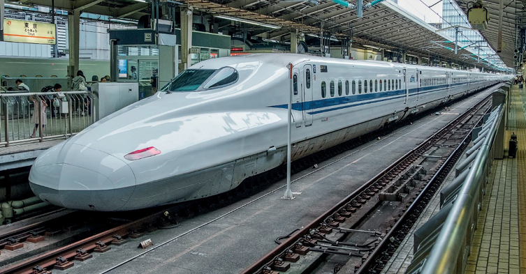 Imagem: Fotografia. Um trem extenso com a parte da frente comprida. Nas laterais há pessoas em plataformas.   Fim da imagem.